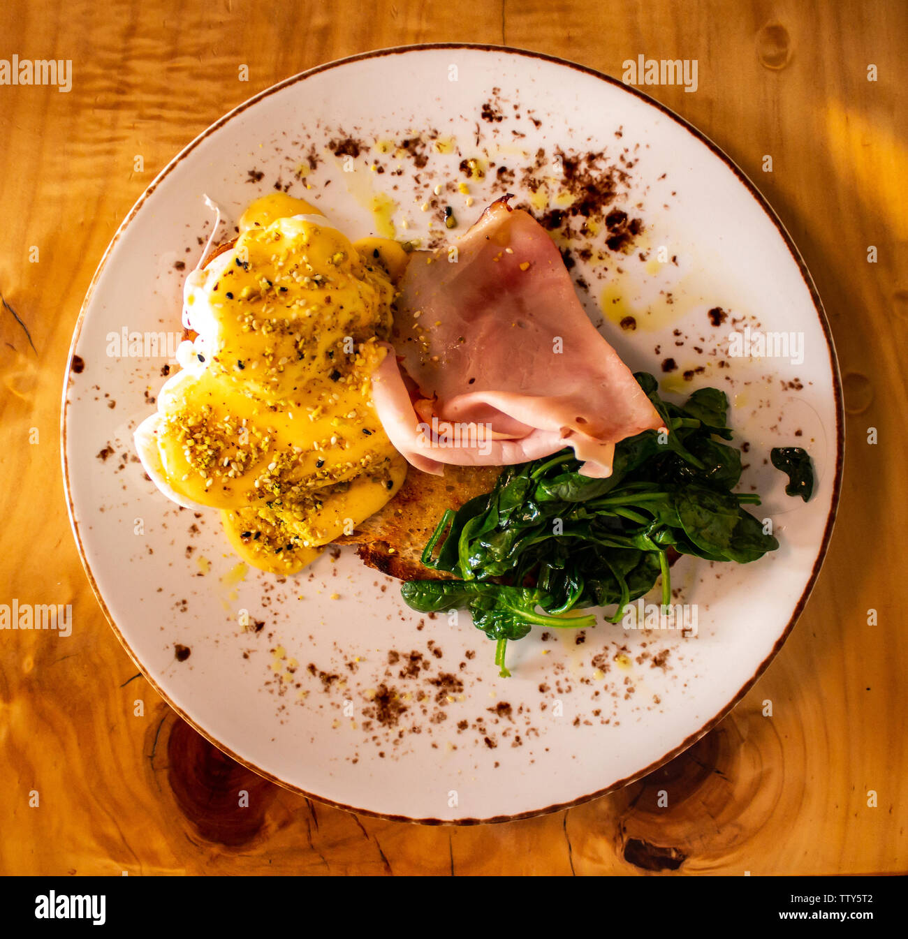 Breakfast meal plate of food comprising eggs Benedict with ham, spinach, toast and garnish set atop wooden table Stock Photo