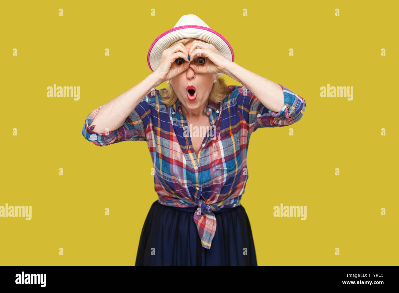 Portrait of funny surprised modern stylish mature woman in casual style with hat standing with binoculars gesture and looking with amazed face. indoor Stock Photo