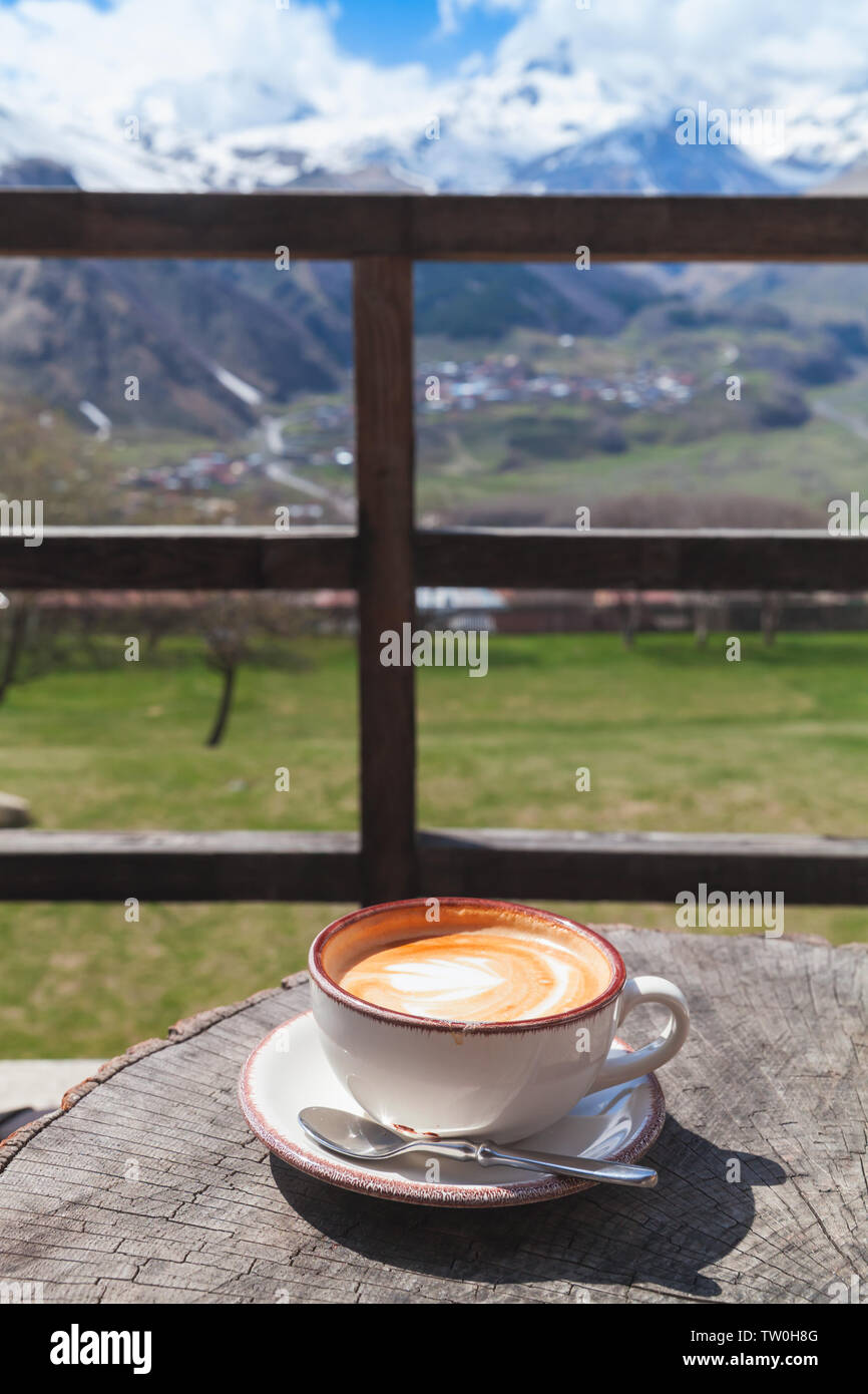 Cappuccino on a balcony. Cup of coffee with milk foam stands on wooden table in morning sunlight with blurred mountain landscape on a background, vert Stock Photo