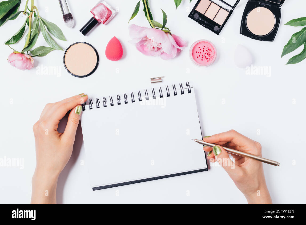 Woman's hands write in blank notepad template next to makeup products and fresh pink flowers peonies on white background, flat lay. Stock Photo