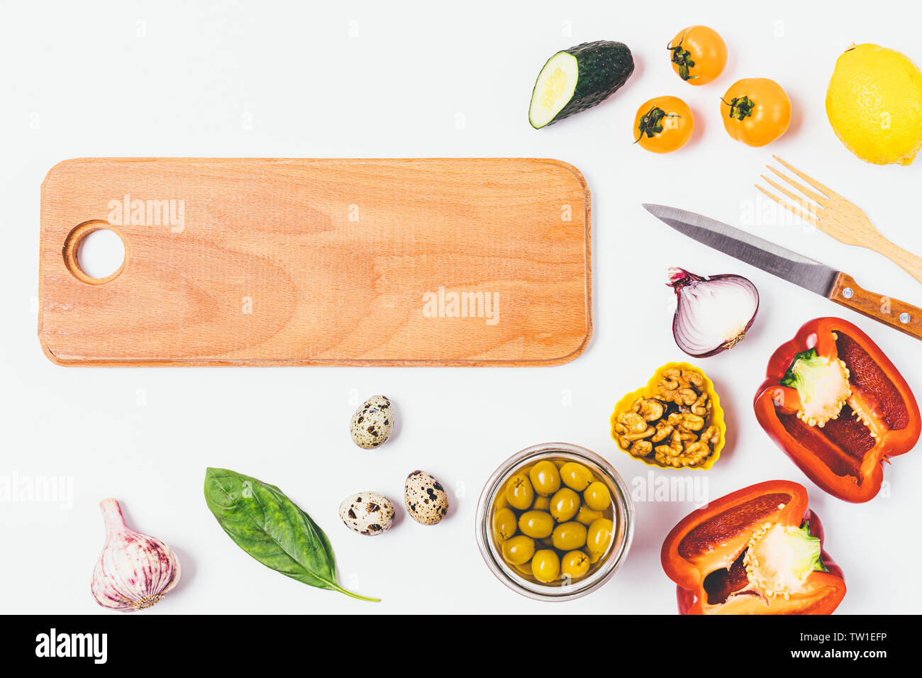 Food background of wooden board next to tomatoes, cucumbers, bell peppers, nuts, greens and quail eggs, flat lay composition on white kitchen table. Stock Photo