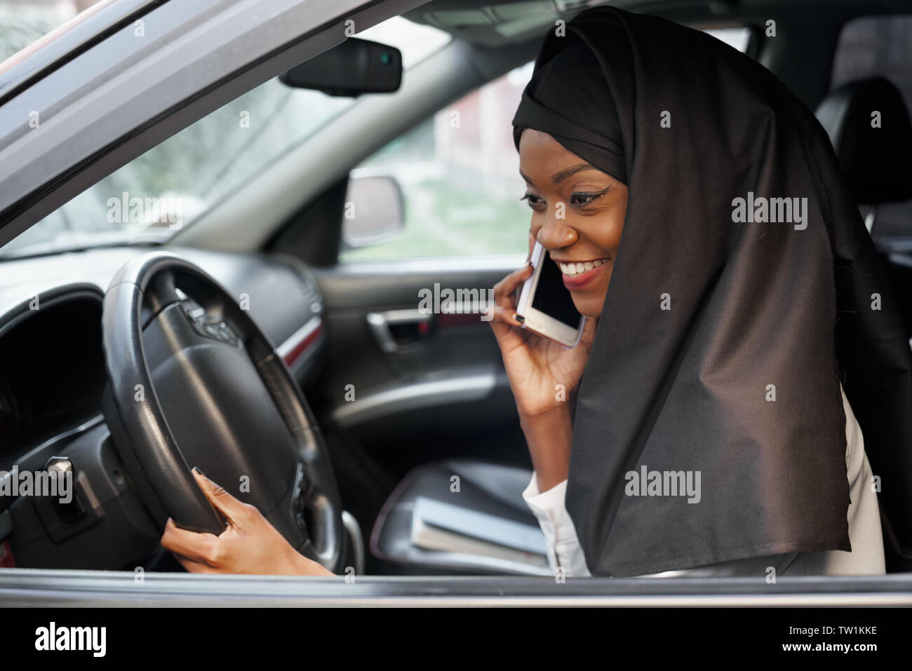 Stylish african muslim woman wearing in black hijab sitting in automobile, talking by mobile phone. Beautiful, young girl holding hand on steering wheel, smiling. Stock Photo