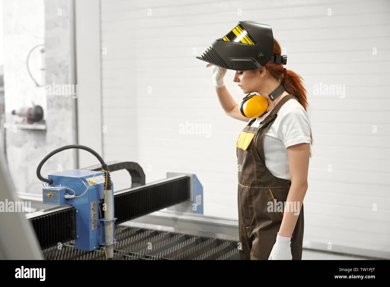 Side view of young female mechanic in protective mask and uniform standing near cnc and controlling process of plasma cutting on metal factory. Concept of technology and machinery. Stock Photo