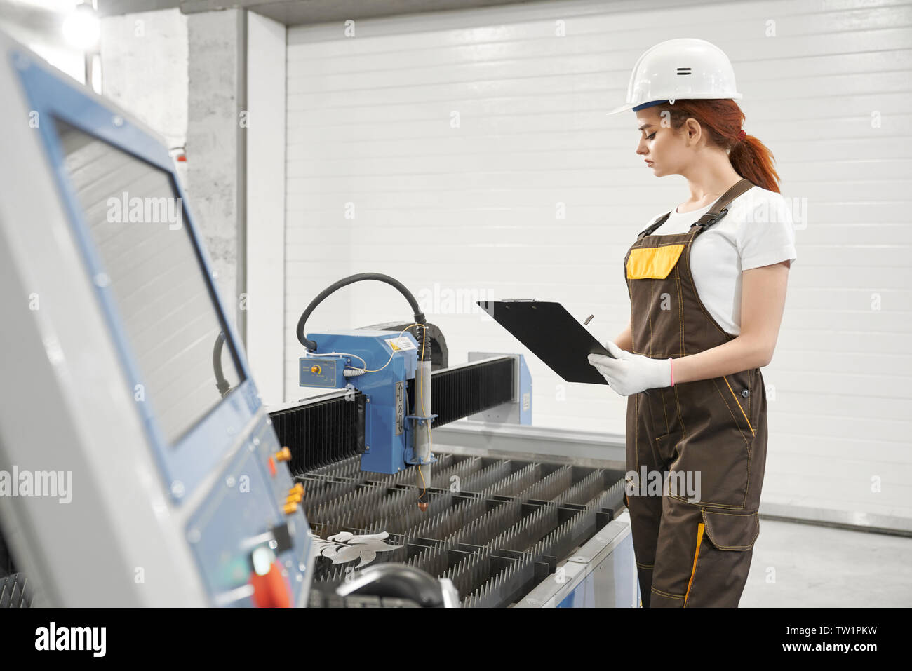 View from side of attractive female engineer in process of controlling plasma cutting device. Young woman in helmet and uniform standing near equipment and writing data. Concept of metalwork. Stock Photo