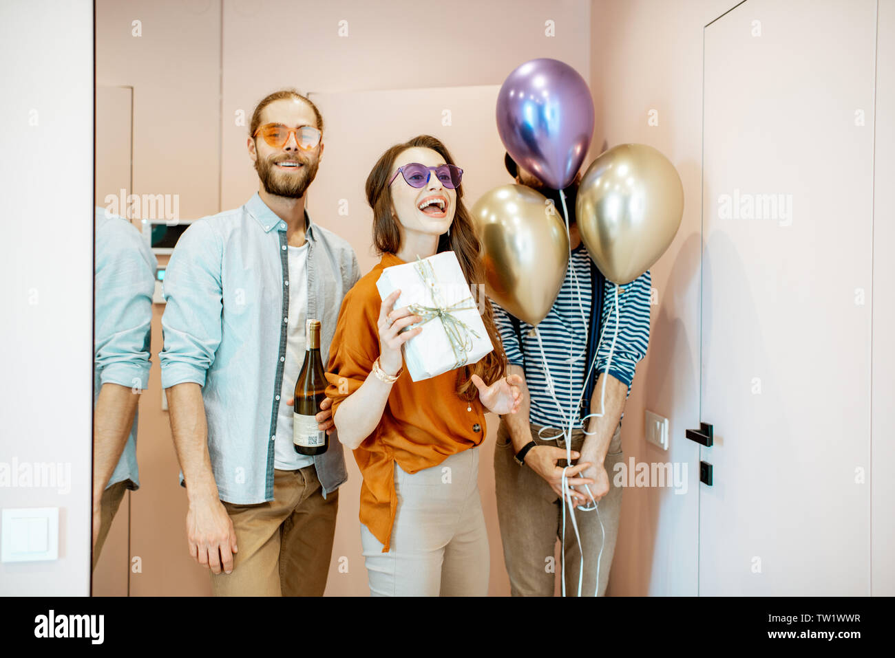 Joyful friends coming with balloons, gifts and drink for the celebration, standing in the apartment hall near the entrance Stock Photo