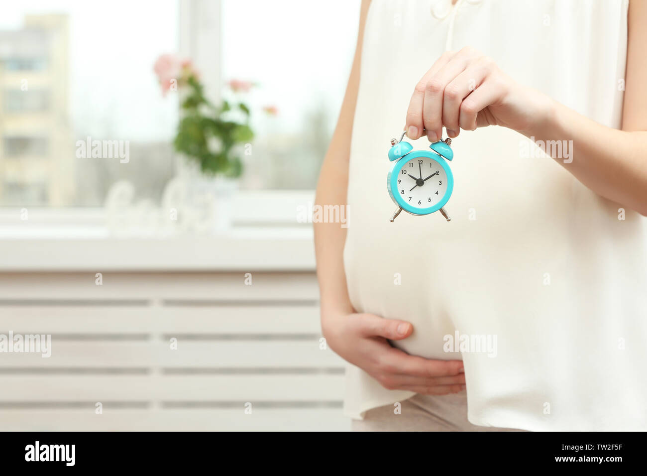 Young pregnant woman holding alarm clock near belly at home Stock Photo