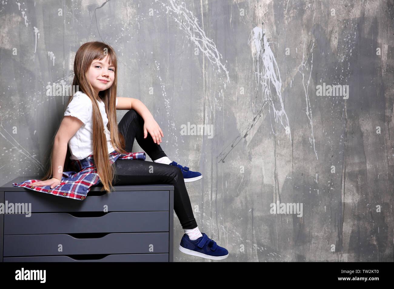 Cute Little Girl Sitting On Chest Of Drawers Against Textured Wall
