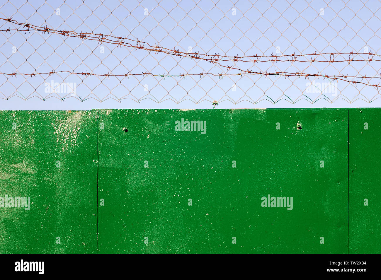 Fence topped with chain-link and barbed wire against clear sky, metal surface roughly painted with green paint Stock Photo