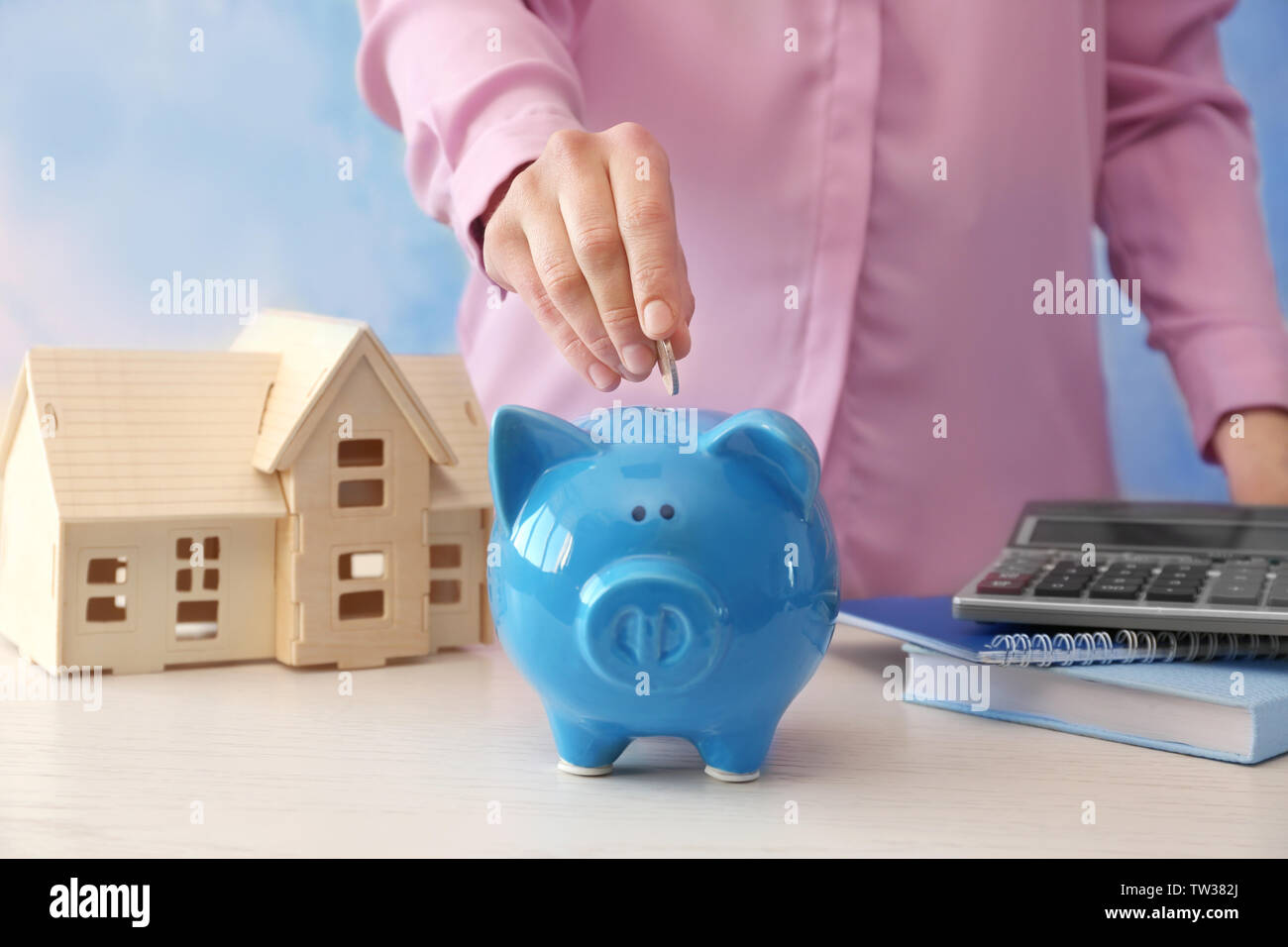 Young woman putting coin into piggy bank on table near house model ...