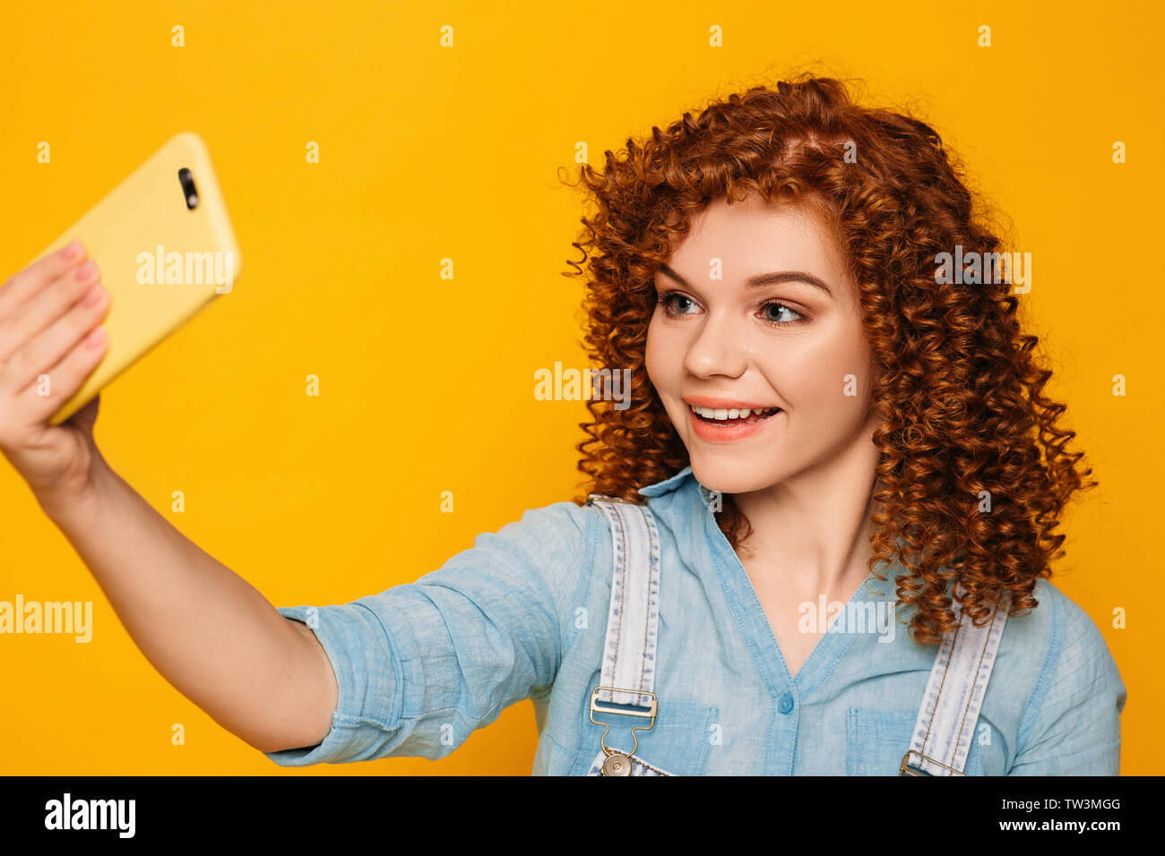 curly haired positive woman making photo on her smartphone on yellow background Stock Photo