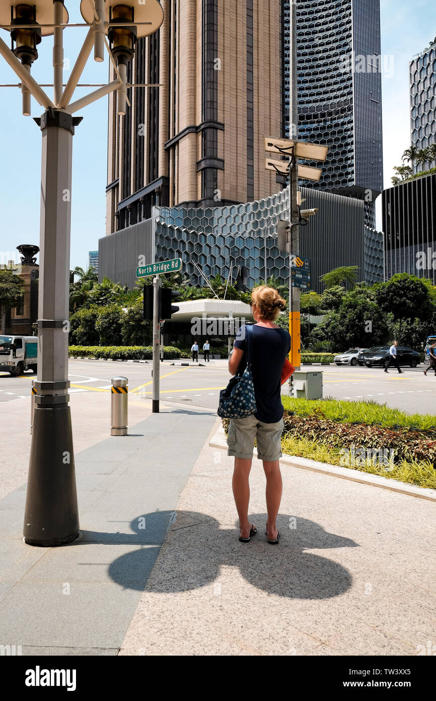 A middle age women shelters under a lamppost shadow to escape the midday sun and heat of Singapore in the Bugis area next to Park View Square Stock Photo