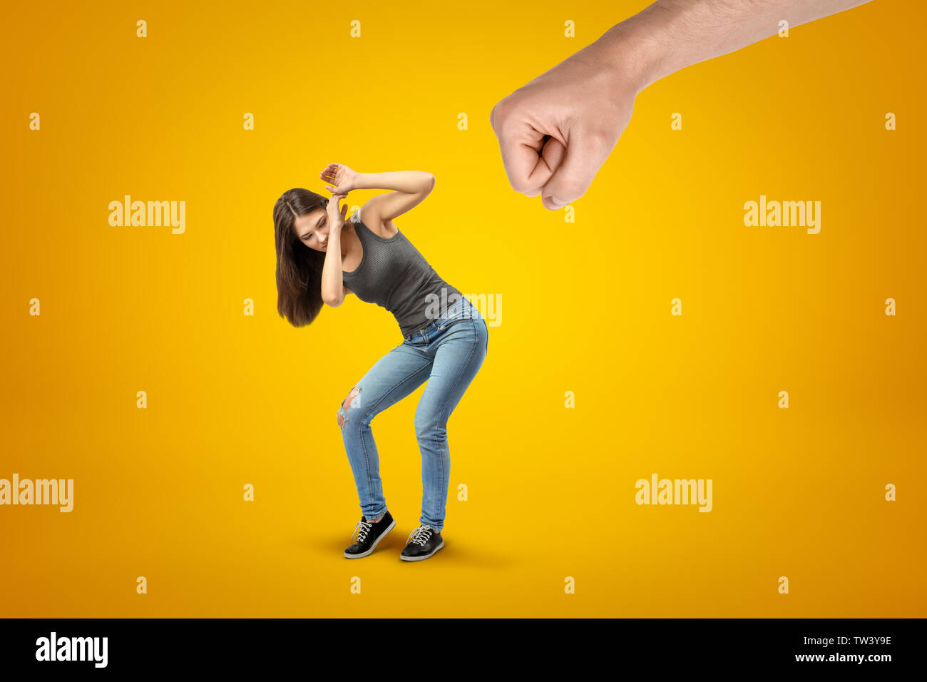 Young brunette girl wearing casual jeans and t-shirt protecting herself from big male stretched fist on yellow background Stock Photo