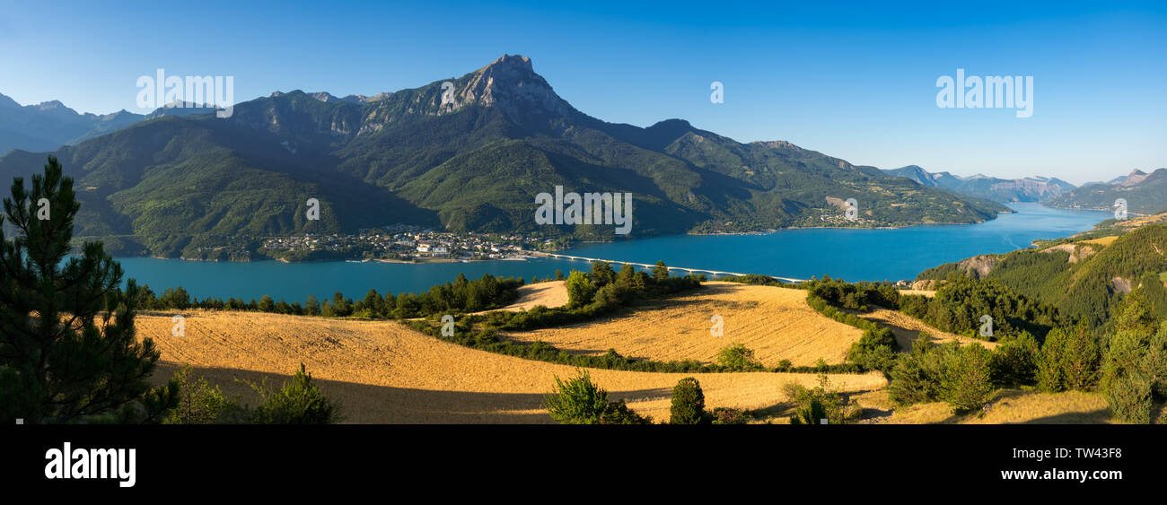 Panoramic of Serre-Poncon lake with the village of Savines-le-Lac and wheatfields in Summer morning light. Hautes-Alpes, France Stock Photo