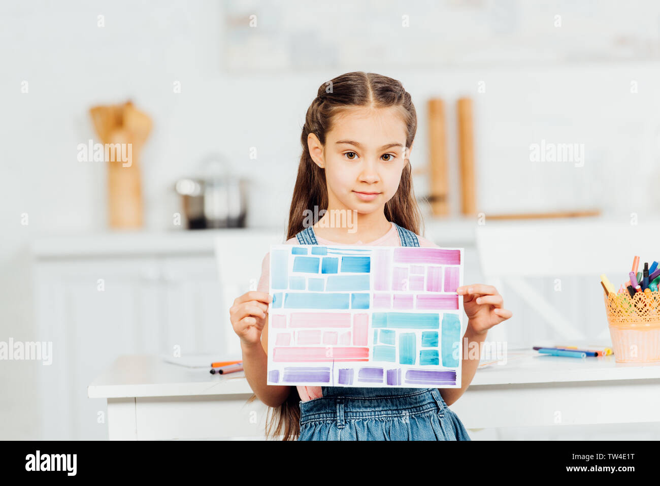 cute child holding paper with colorful stripes while standing near table at home Stock Photo