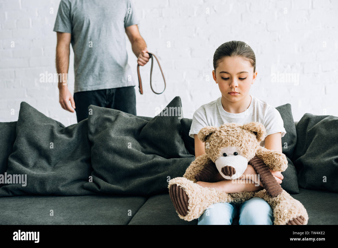 partial view of abusive father with belt and sad daughter with teddy bear sitting on sofa Stock Photo
