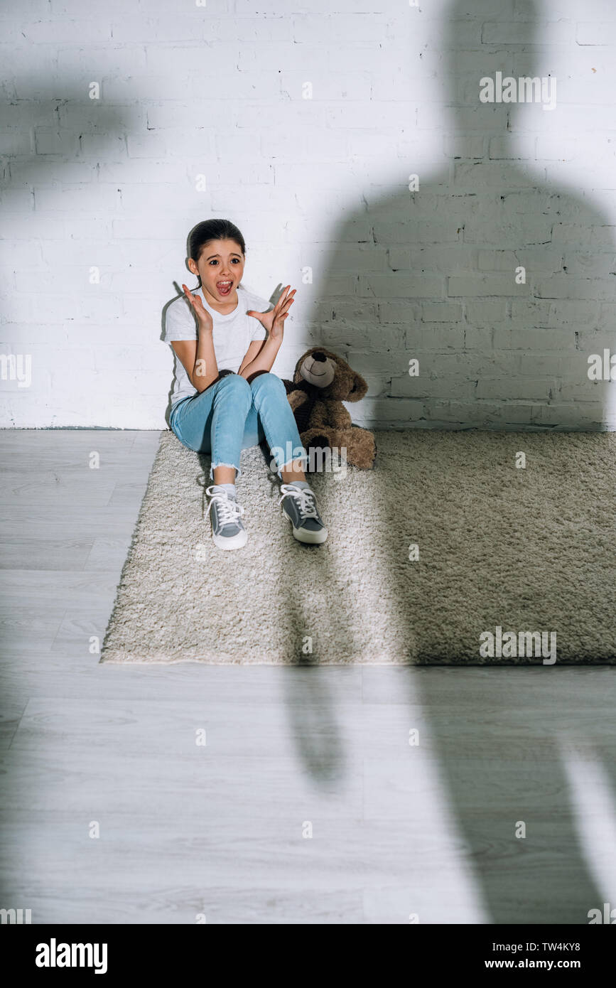 big silhouette and scared kid with teddy bear screaming while sitting on carpet Stock Photo