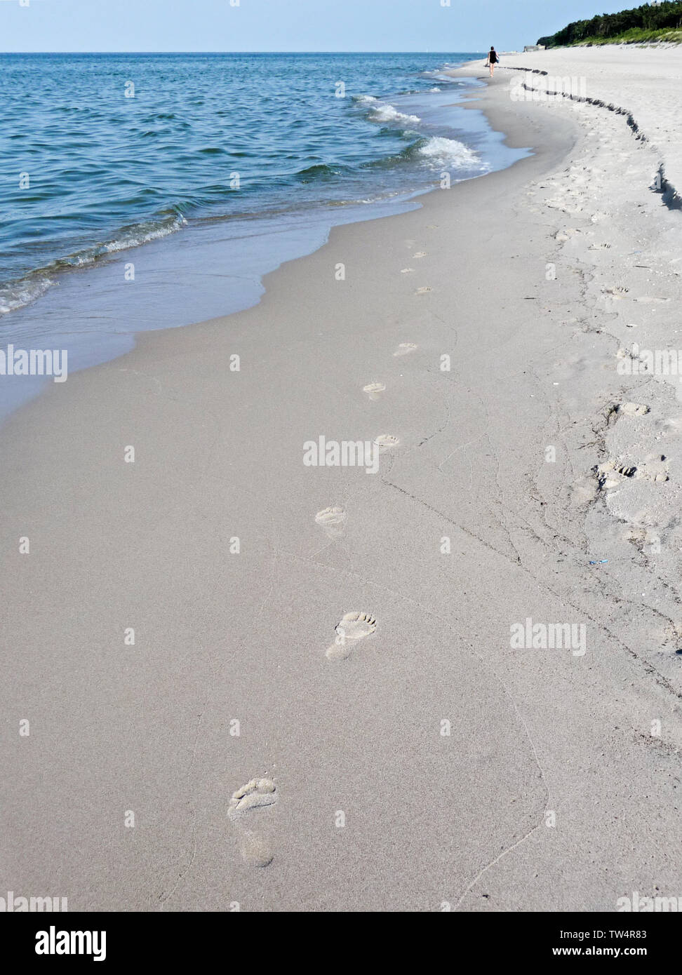 Footprints on beach sand vanishing in sea waves,  in the distance single human figure walking along the seacoast Stock Photo