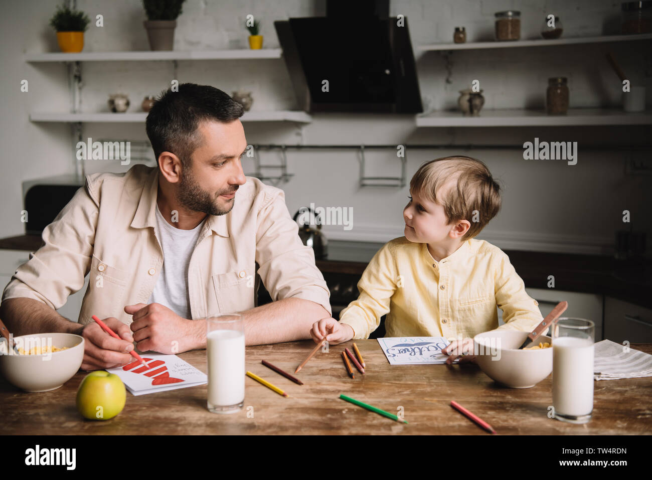 father and son drawing while sitting at kitchen table with served breakfast Stock Photo