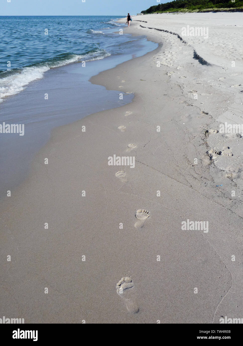 Footprints on beach sand vanishing in sea waves,  in the distance single human figure walking along the seacoast Stock Photo