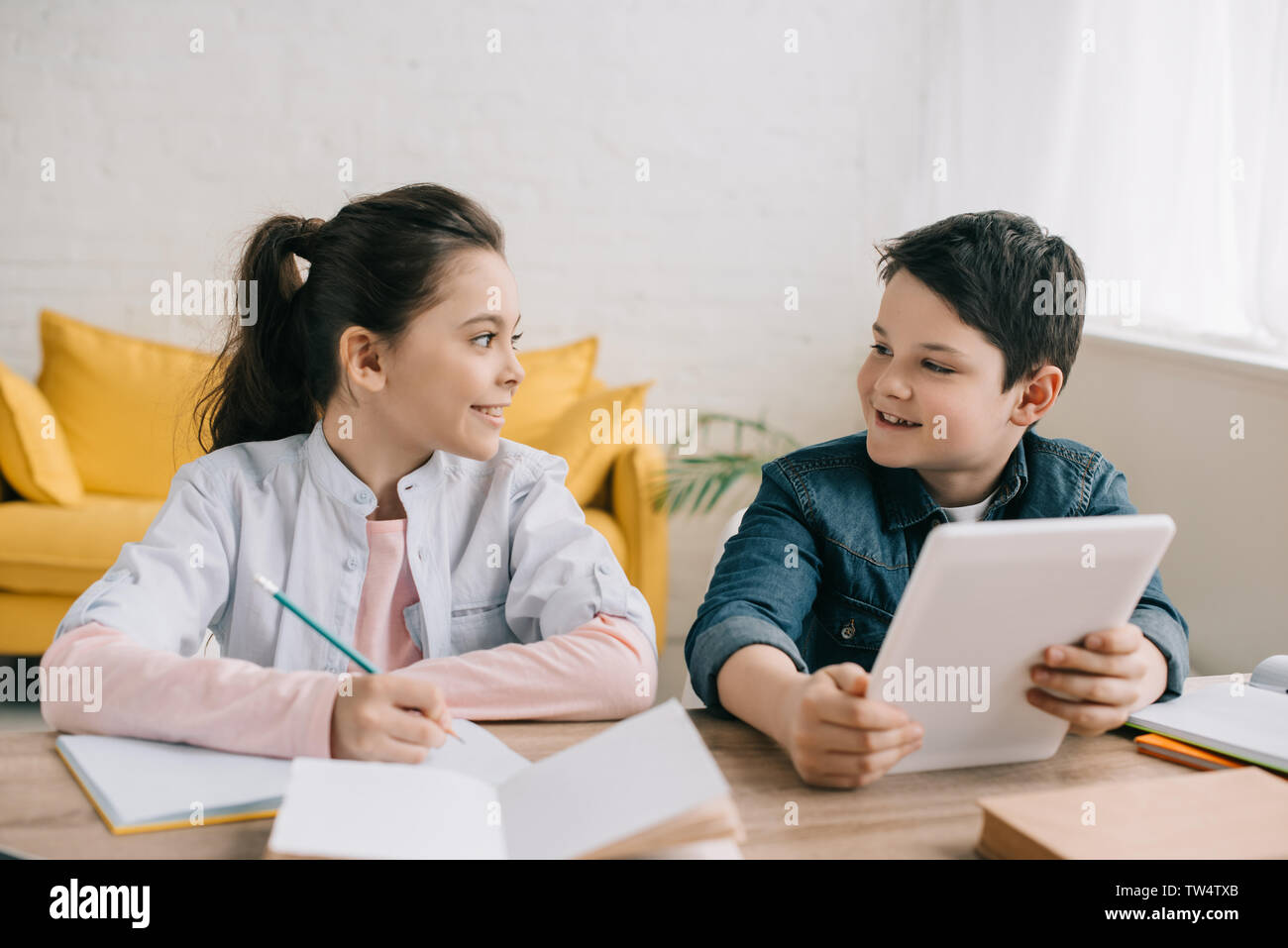 smiling boy using digital tablet while sitting at desk near sister writing in notebook Stock Photo