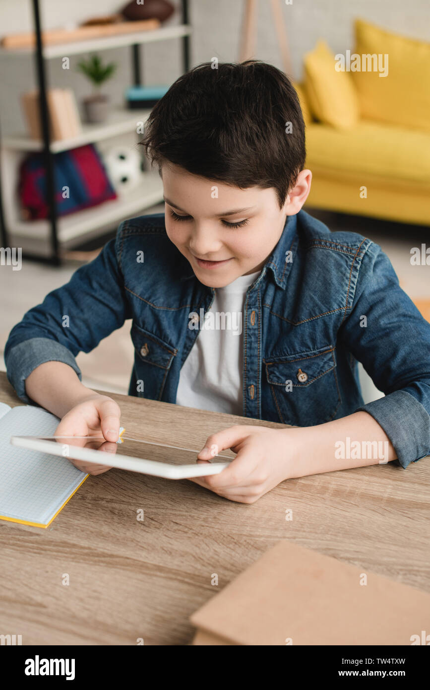 cute smiling boy using digital tablet while sitting at wooden desk at ...