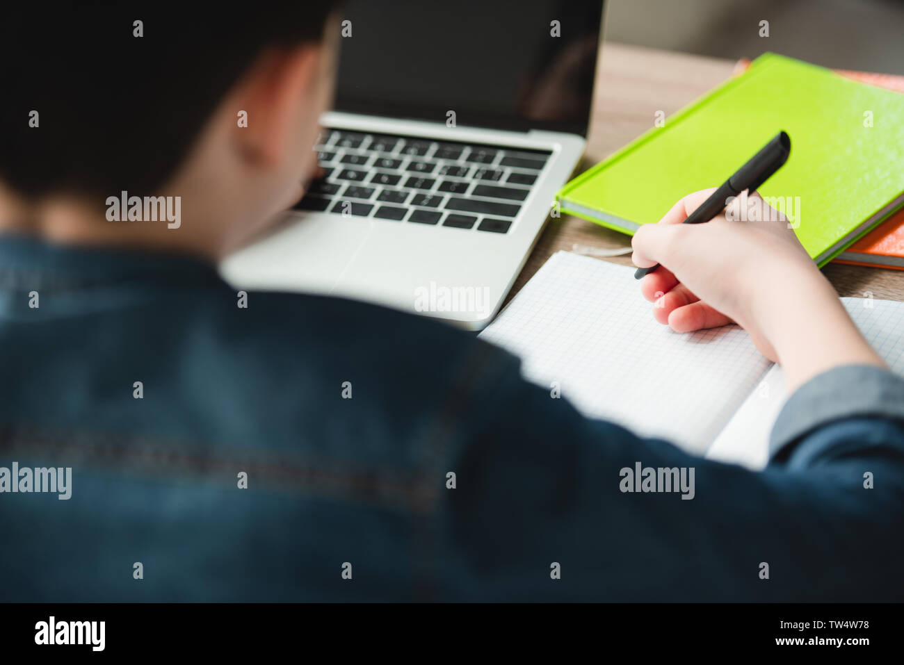 back view of boy writing in notebook and using laptop while doing homework Stock Photo