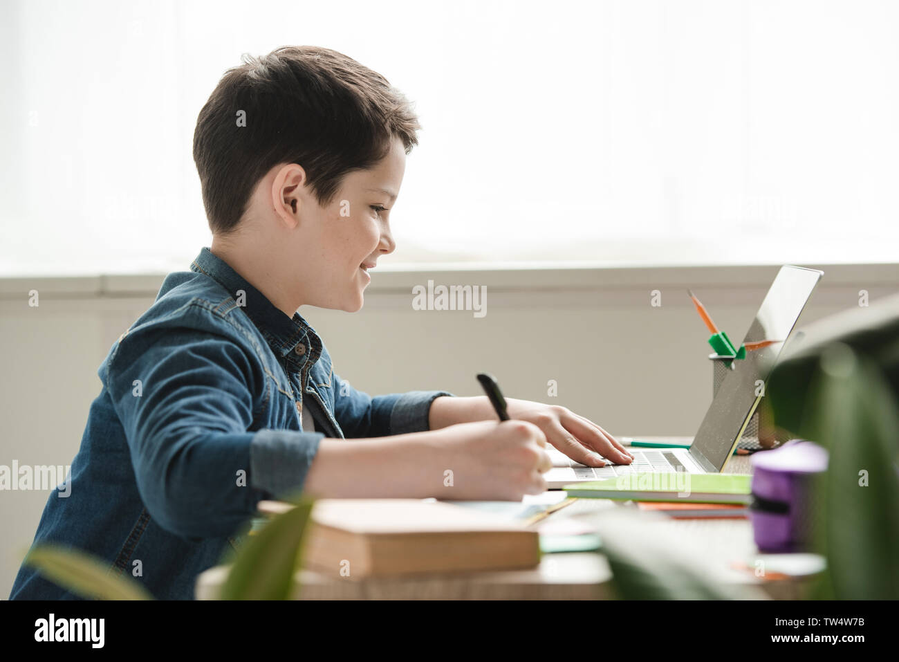 smiling boy writing in notebook and using laptop while doing schoolwork at home Stock Photo