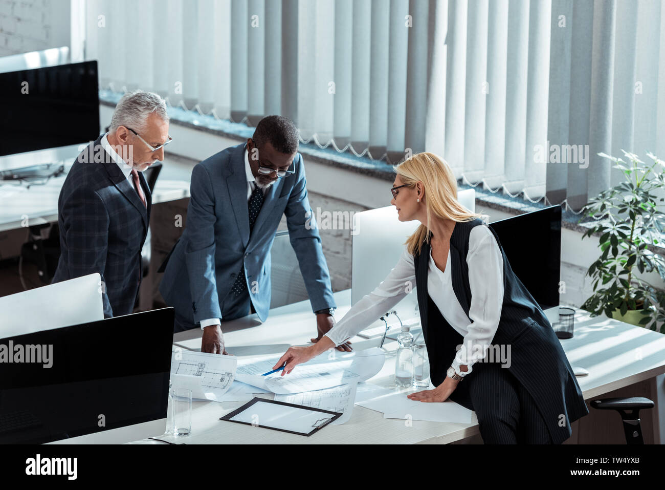 overhead view of blonde businesswoman in glasses holding pen near blueprints and multicultural businessmen Stock Photo