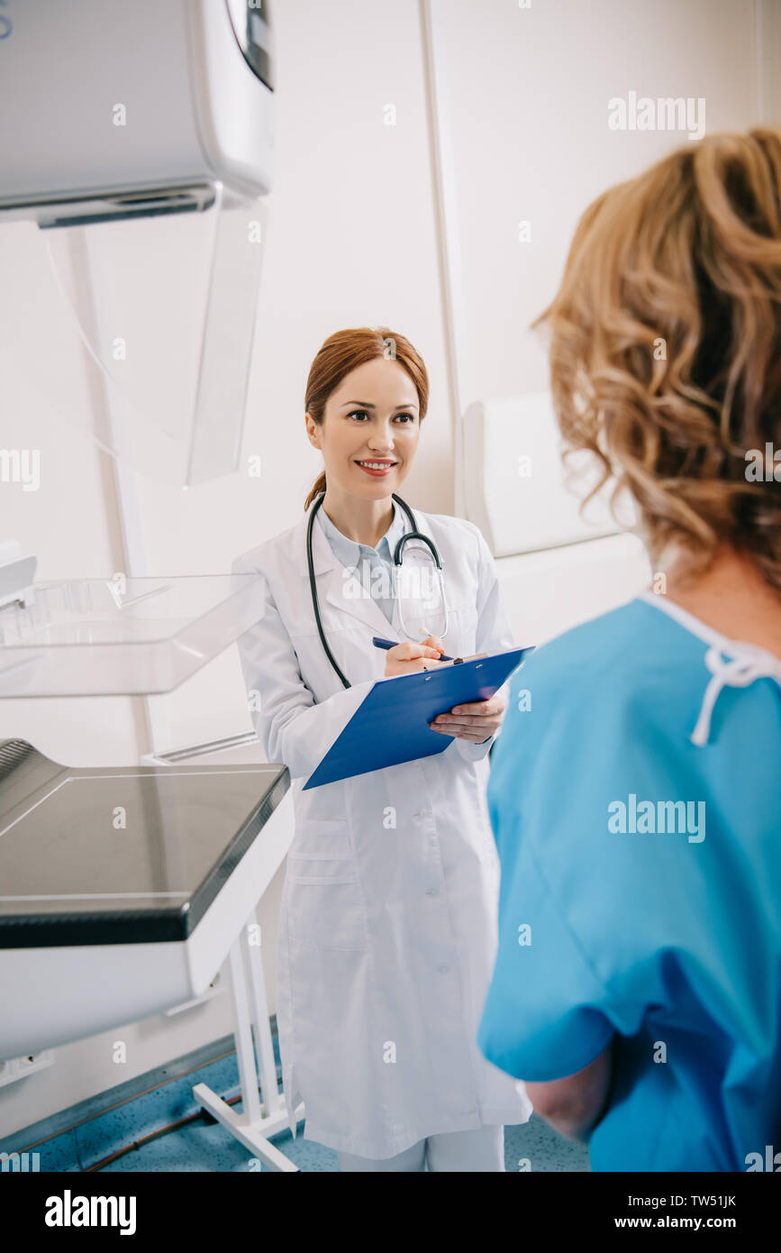 selective focus of smiling radiographer writing on clipboard near patient in clinic Stock Photo