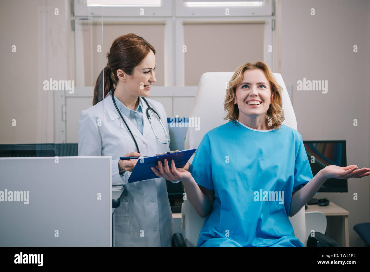 happy patient gesturing while sitting in armchair near smiling doctor holding clipboard Stock