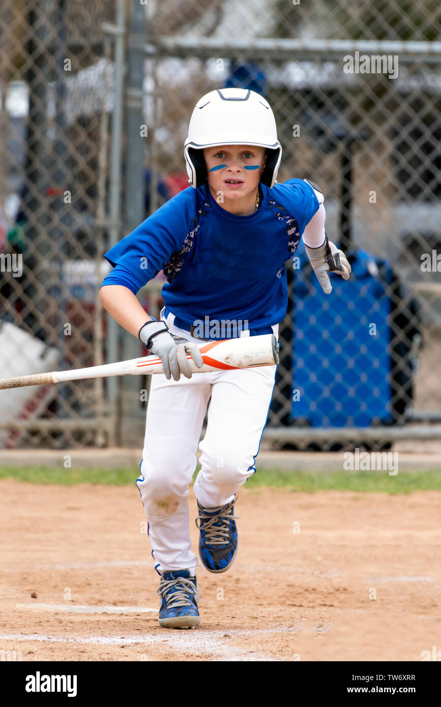 Youth baseball player in blue uniform and white helmet running up the base line with bat in hand. Stock Photo