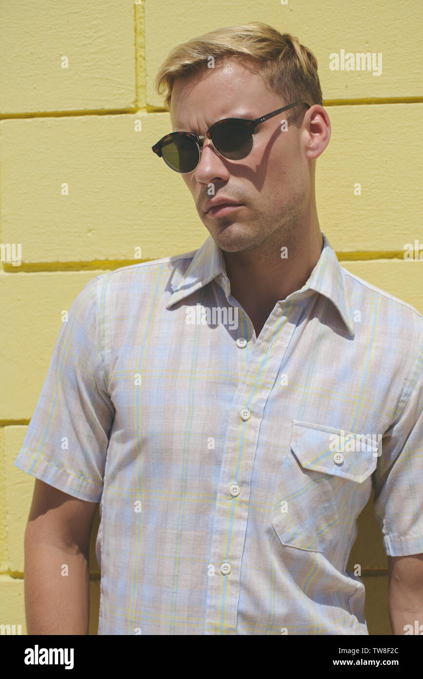 A blonde male model wearing a western plaid shirt and dark sunglasses, posing against a yellow wall standing outdoor. Stock Photo