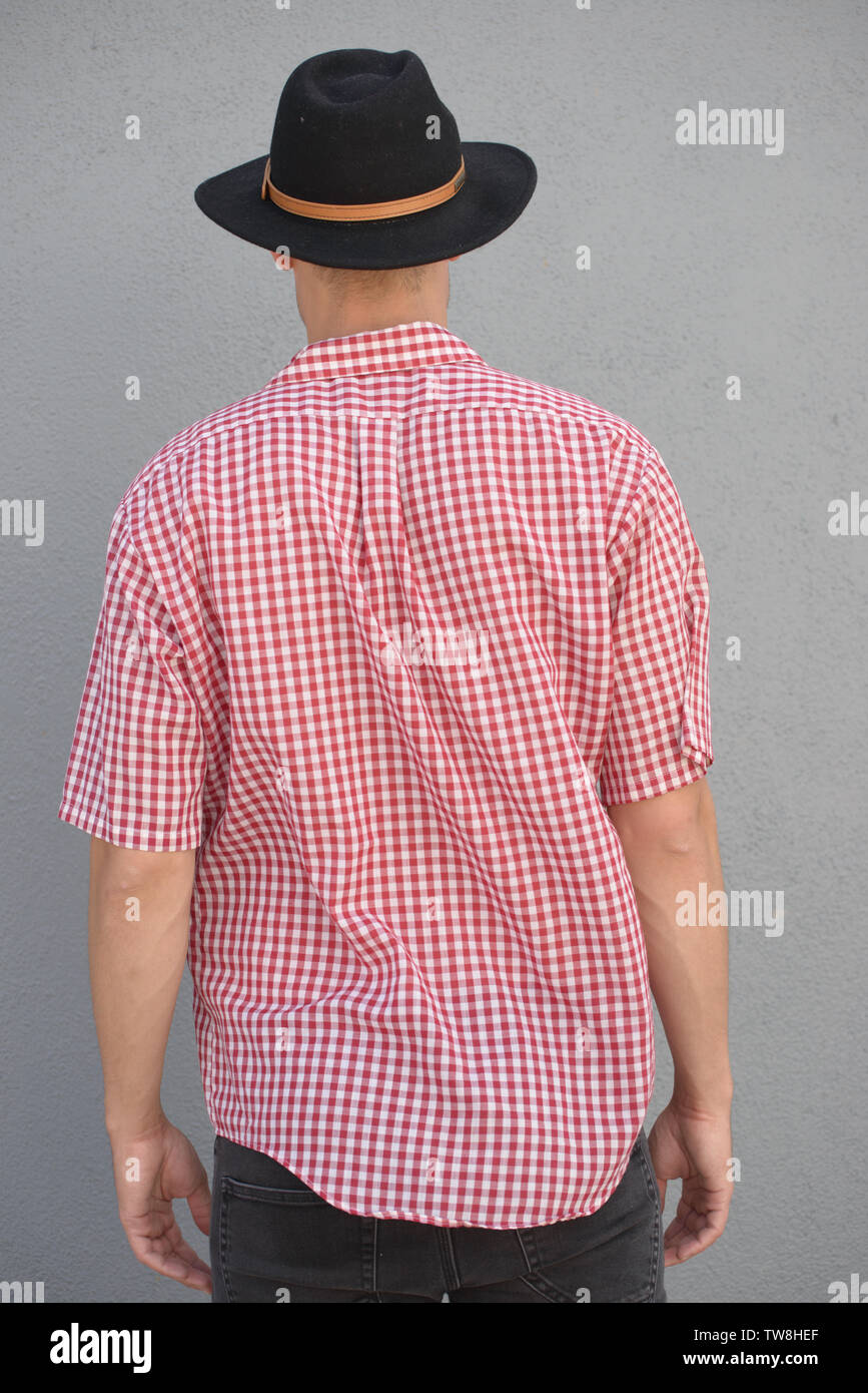 A blonde male model wearing a western plaid shirt, with fedora hat, posing with his back facing camera, standing against a wall standing outdoor. Stock Photo