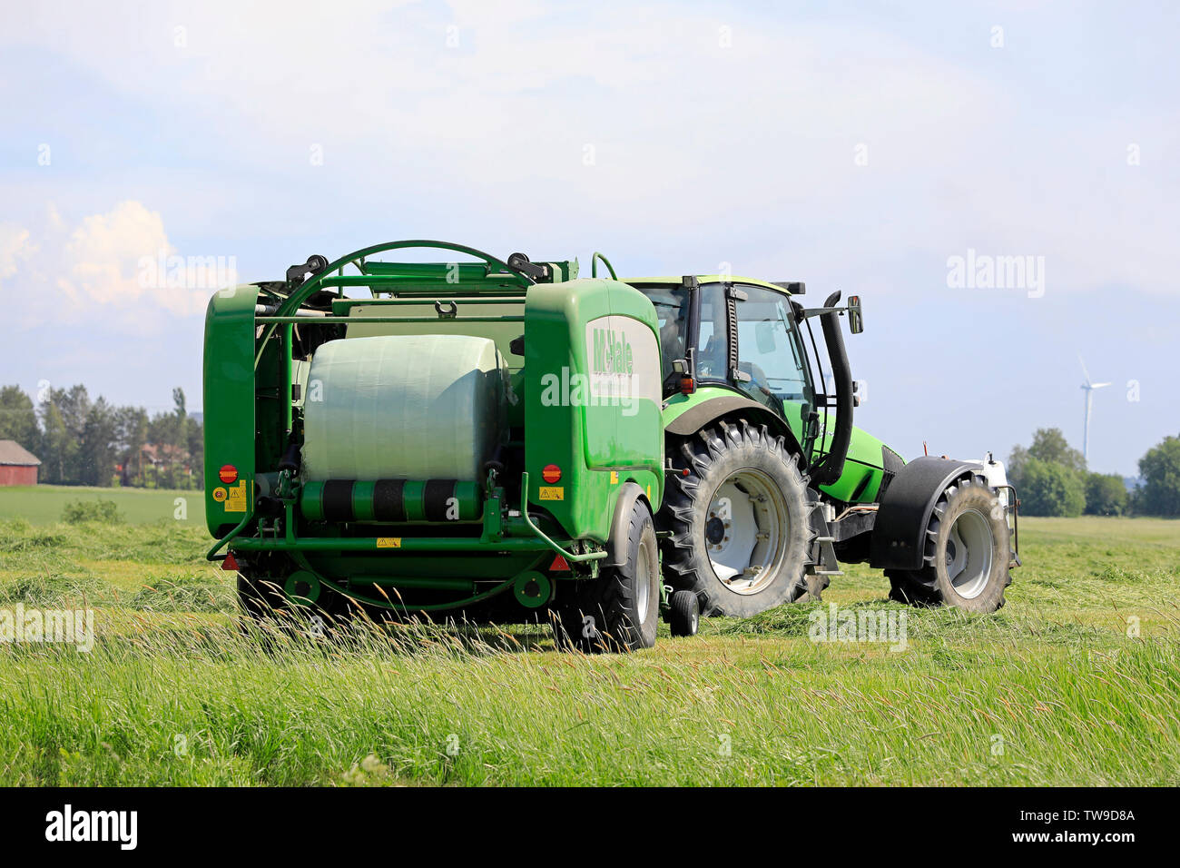 Salo, Finland. June 15, 2019. Deutz-Fahr tractor and McHale 3 plus baler baling silage in green plastic sheet in hay field on a sunny day of summer. Stock Photo