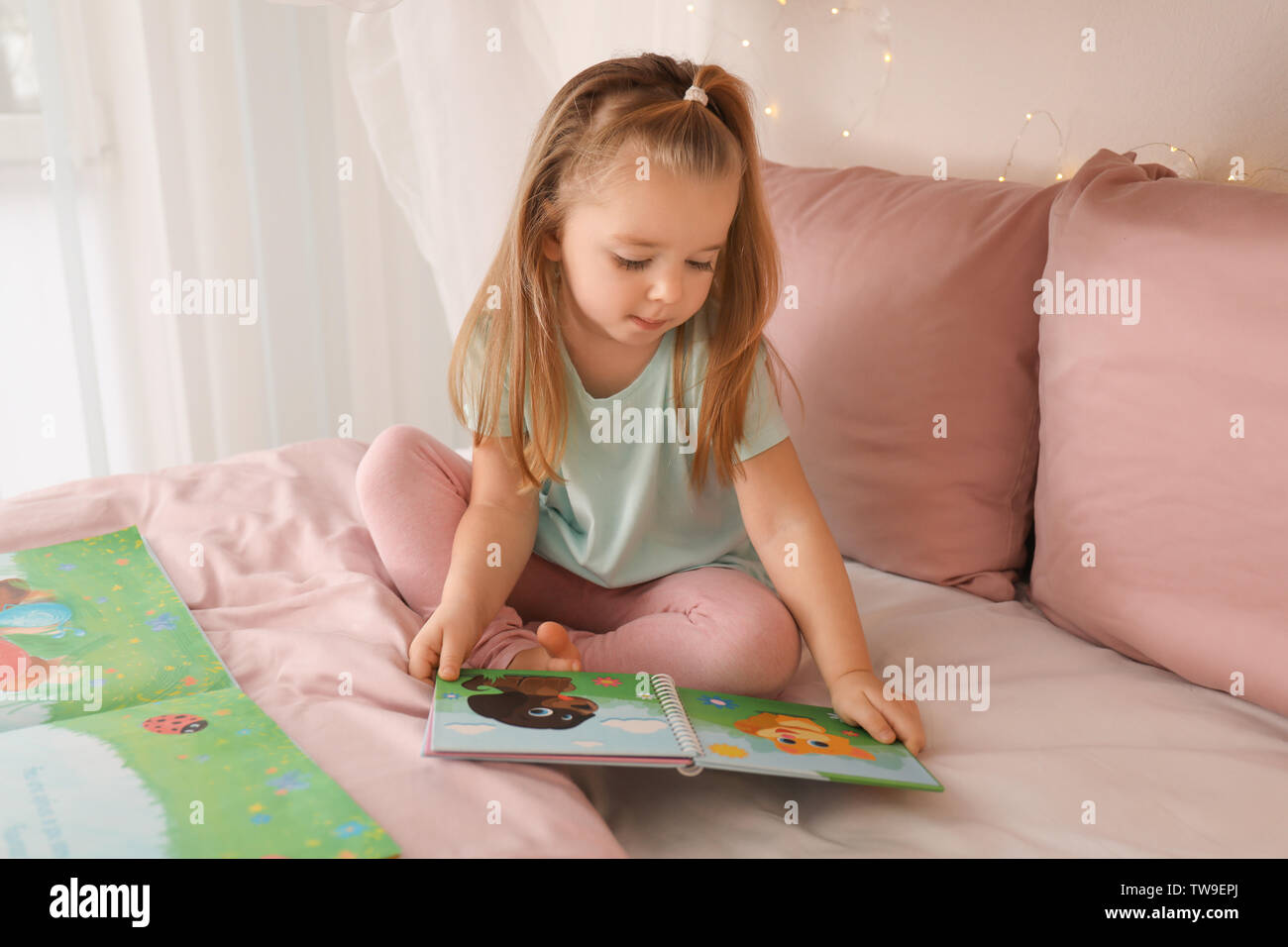 Cute little girl reading book while sitting on bed at home Stock Photo ...