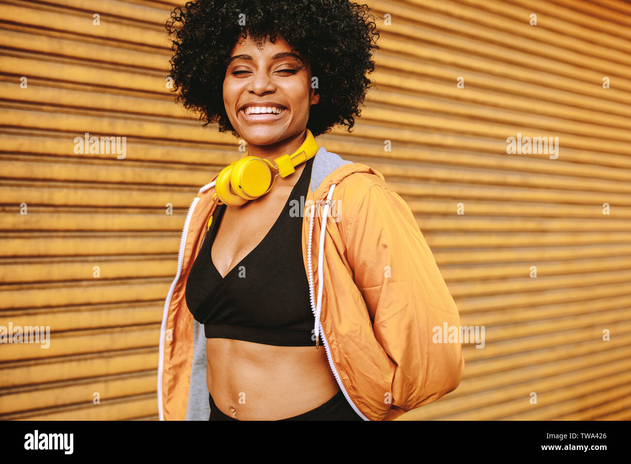 Sporty young woman standing outdoors in the city and smiling. Female athlete with headphones resting after workout. Stock Photo