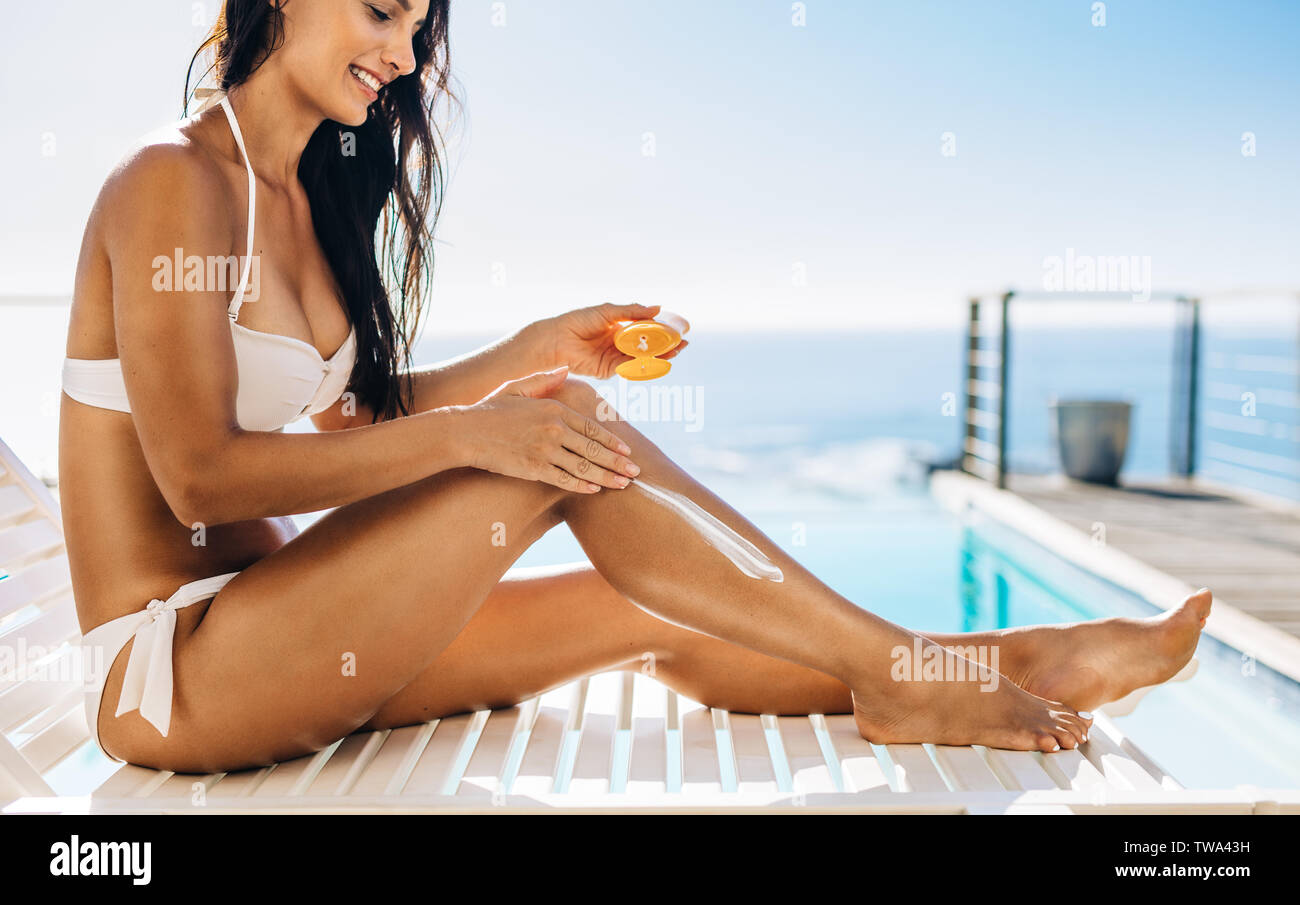 Happy young woman sitting on deck chair by the swimming pool applying sun cream onto her legs. Female model sunbathing at the poolside. Stock Photo