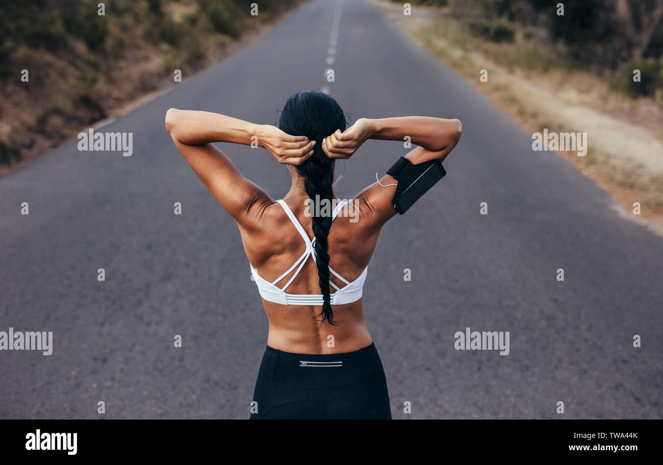 Rear view shot of muscular woman tying her hair and getting ready for a sprint. Fitness woman before a run on empty road. Stock Photo