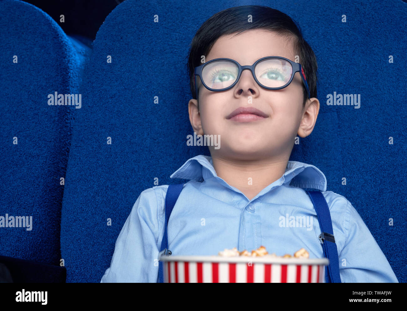 Front view of funny little boy sitting in cinema and watching excited film.  Young spectator wearing glasses and white shirt smiling and relaxing on  weekend. Concept of entertainment and enjoyment Stock Photo -