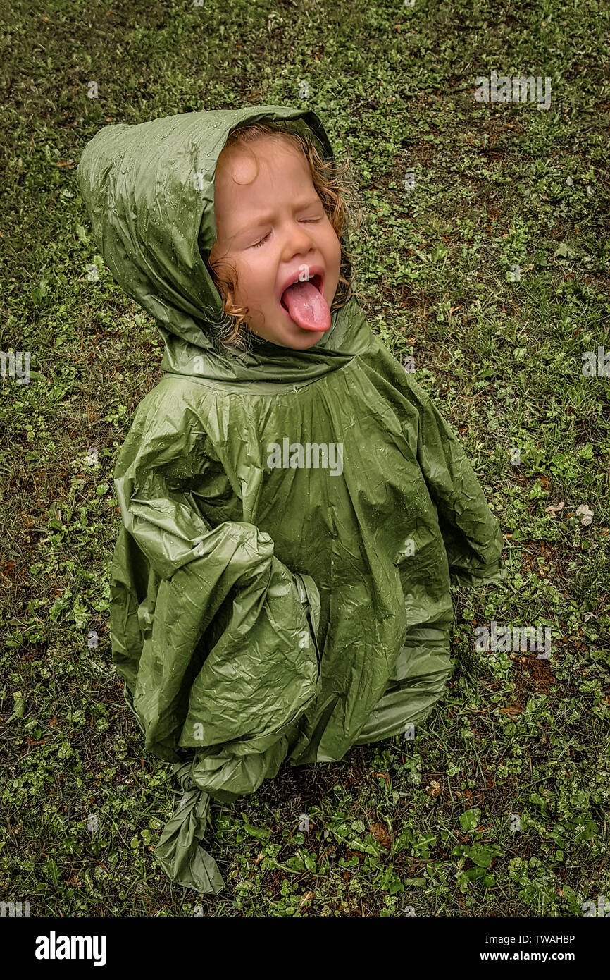 Little toddler girl, playing in rain, trying to taste it, with eyes closed and tongue out, with green rain coat, on a green grass Stock Photo
