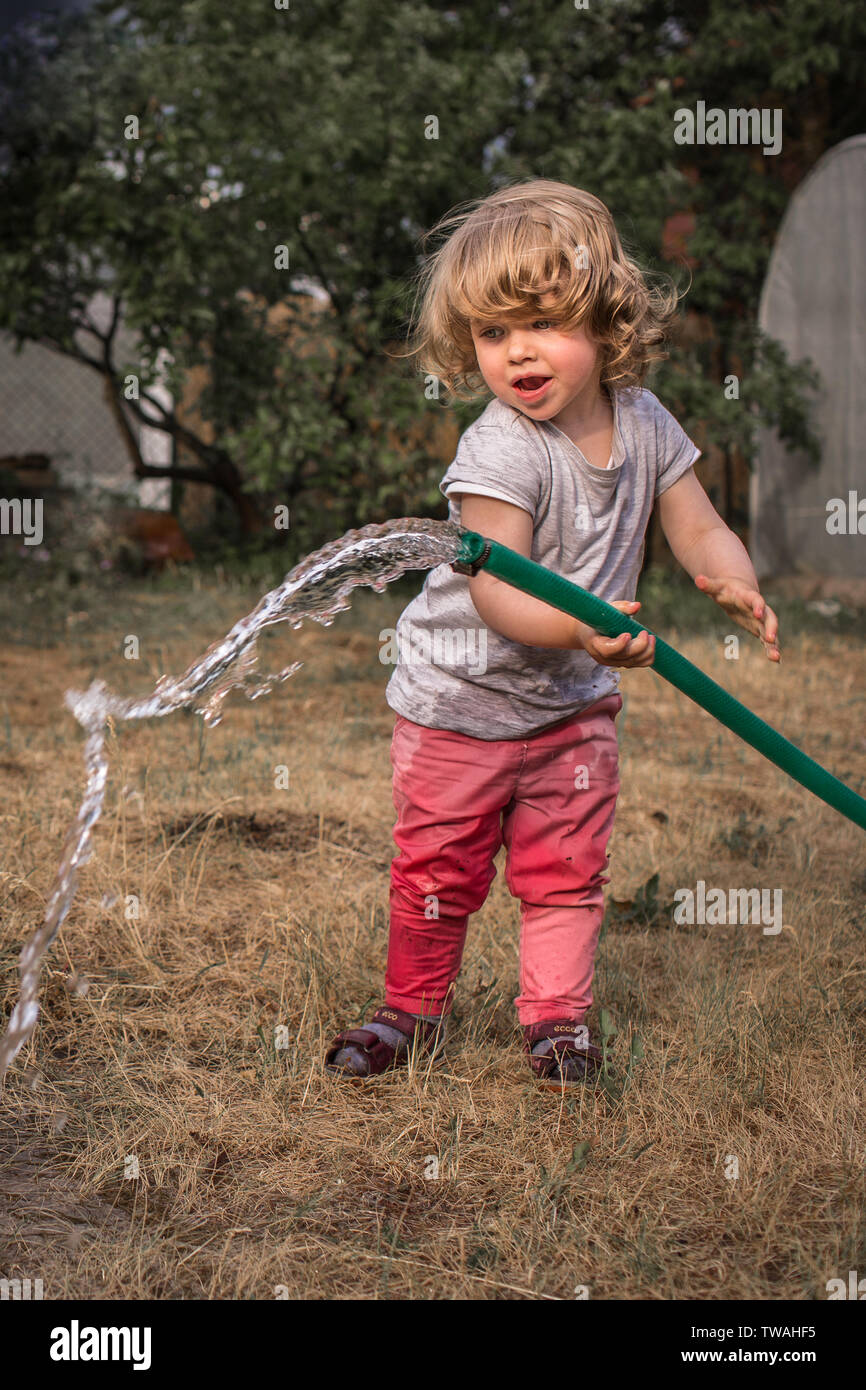 Little, cute girl with curly hair enjoying summer fun, splashing water out of garden hose Stock Photo