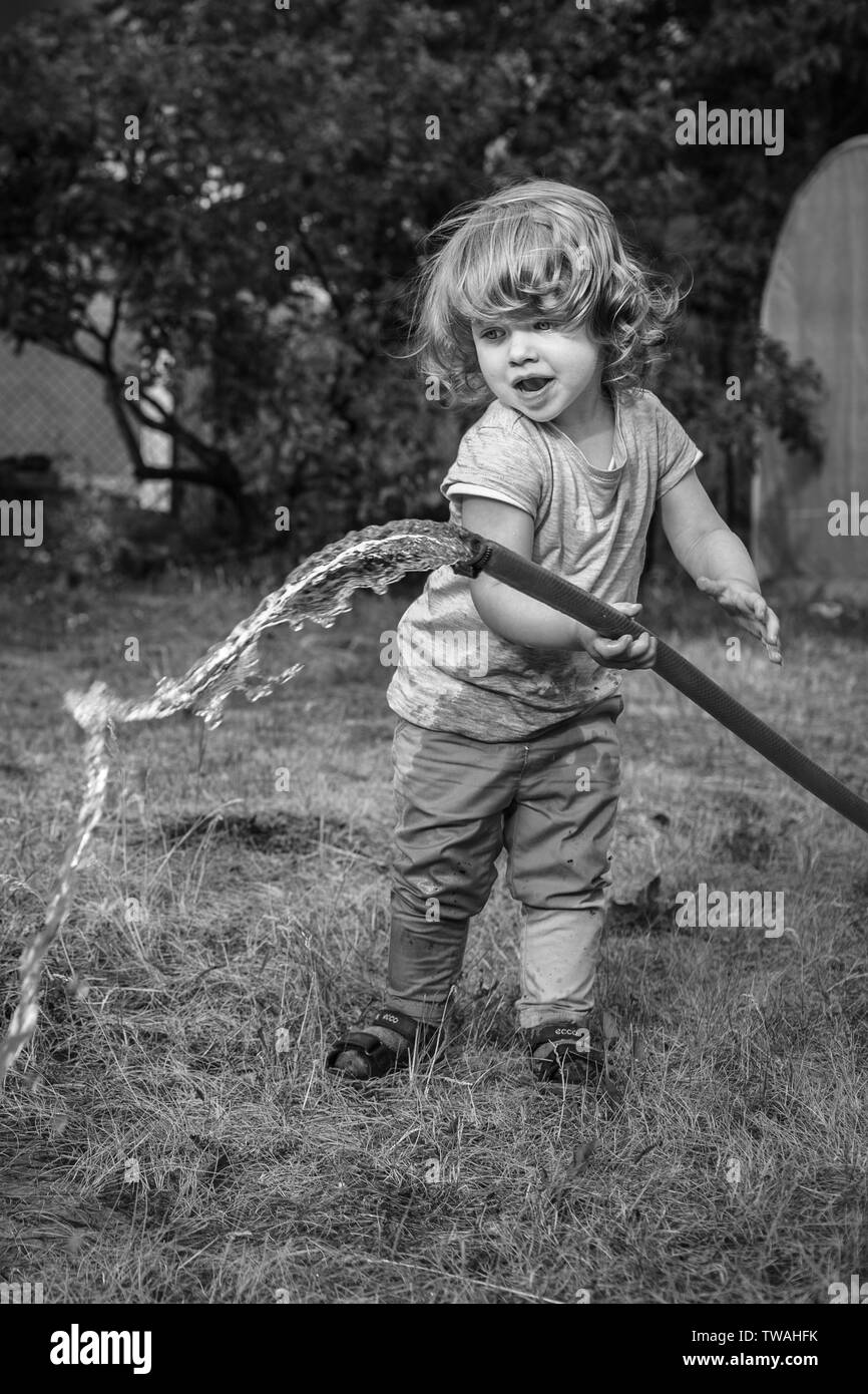 Black and white photo of little, cute girl with curly hair enjoying summer fun, splashing water out of garden hose Stock Photo