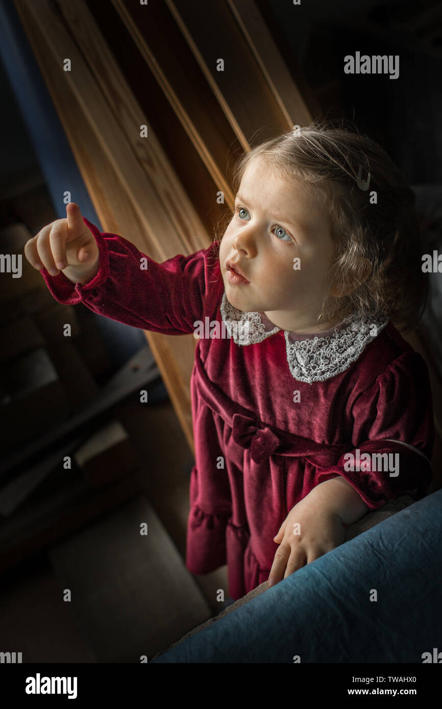 Little, beautiful girl looking up and pointing finger, wearing red velvet Christmas dress with white lace collar Stock Photo
