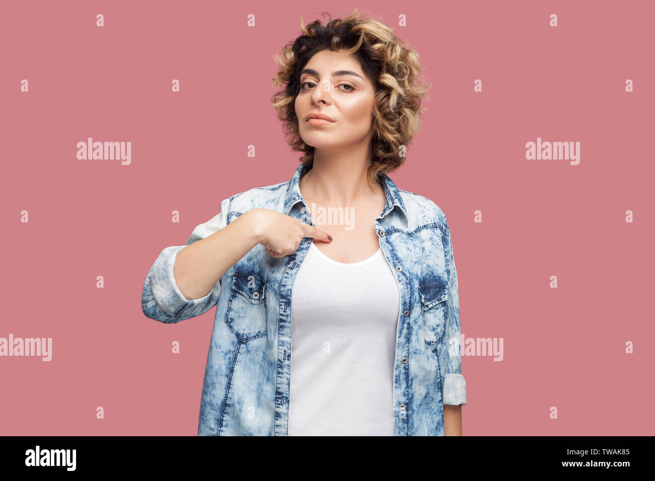 This is me. Portrait of serious young woman with curly hairstyle in casual blue shirt standing, pointing herself with proud face and looking at camera Stock Photo