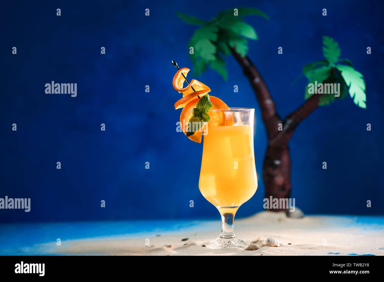 Decorated yellow tropical cocktail in a tulip glass with oranges on a skewer. Papercraft palm trees on a blue background with copy space. Stock Photo