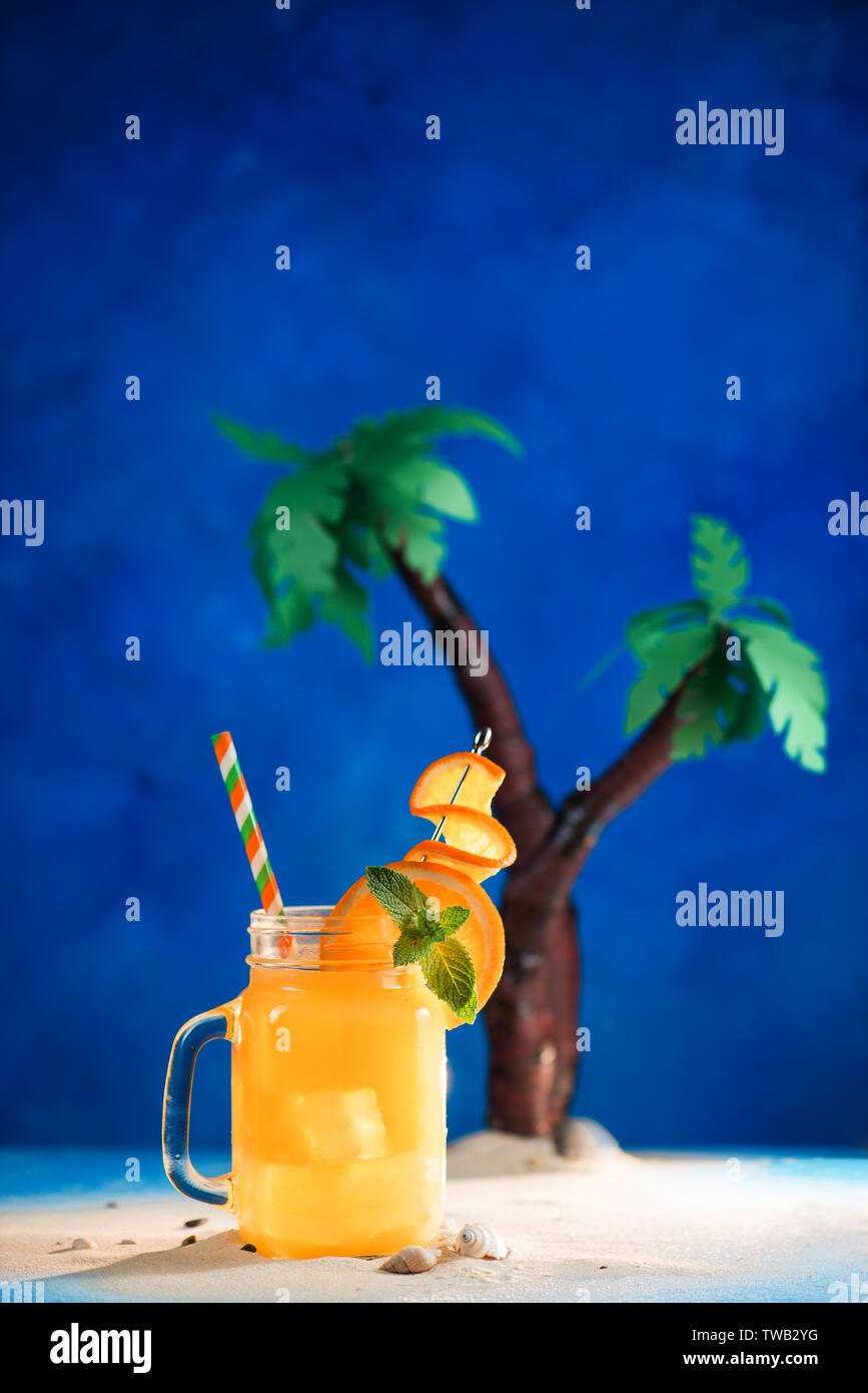 Decorated tropical cocktail in a glass jar on a blue background with copy space. Papercraft palm trees with yellow juice. Stock Photo