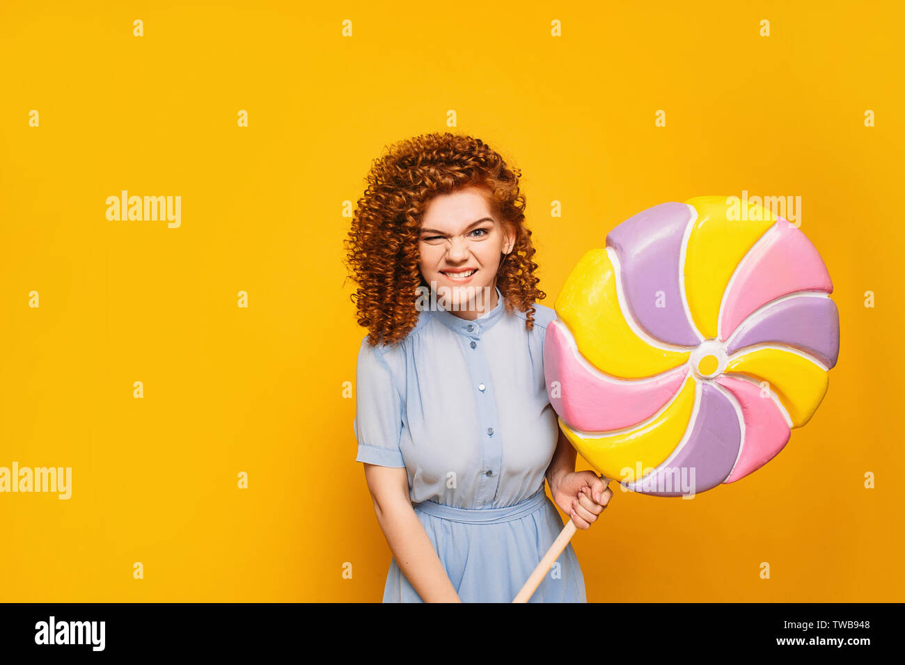 curly red-haired positive woman wearing blue dress with sweet big lollipop on yellow background Stock Photo