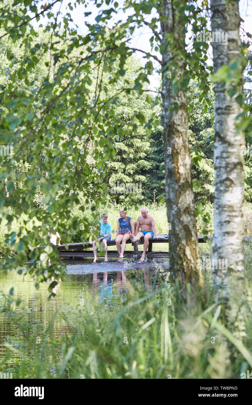 Family spending time together sitting on a bridge over a lake, among the trees, close to nature, during summer vacations. Candid people, real moments, Stock Photo