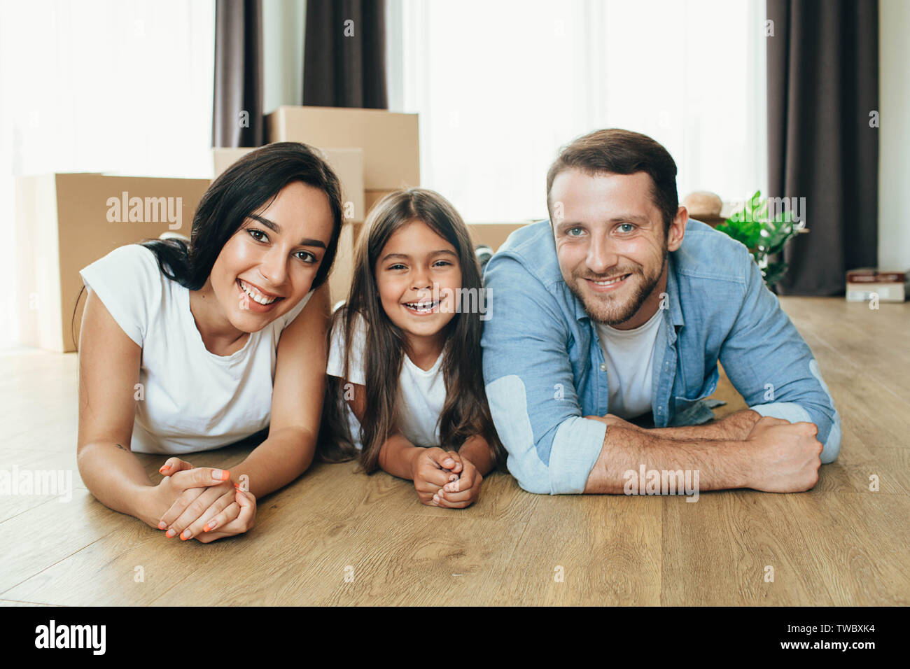 Happy family moving in new house. Mother, father and daughter together smiling lying down on floor with boxes on background Stock Photo