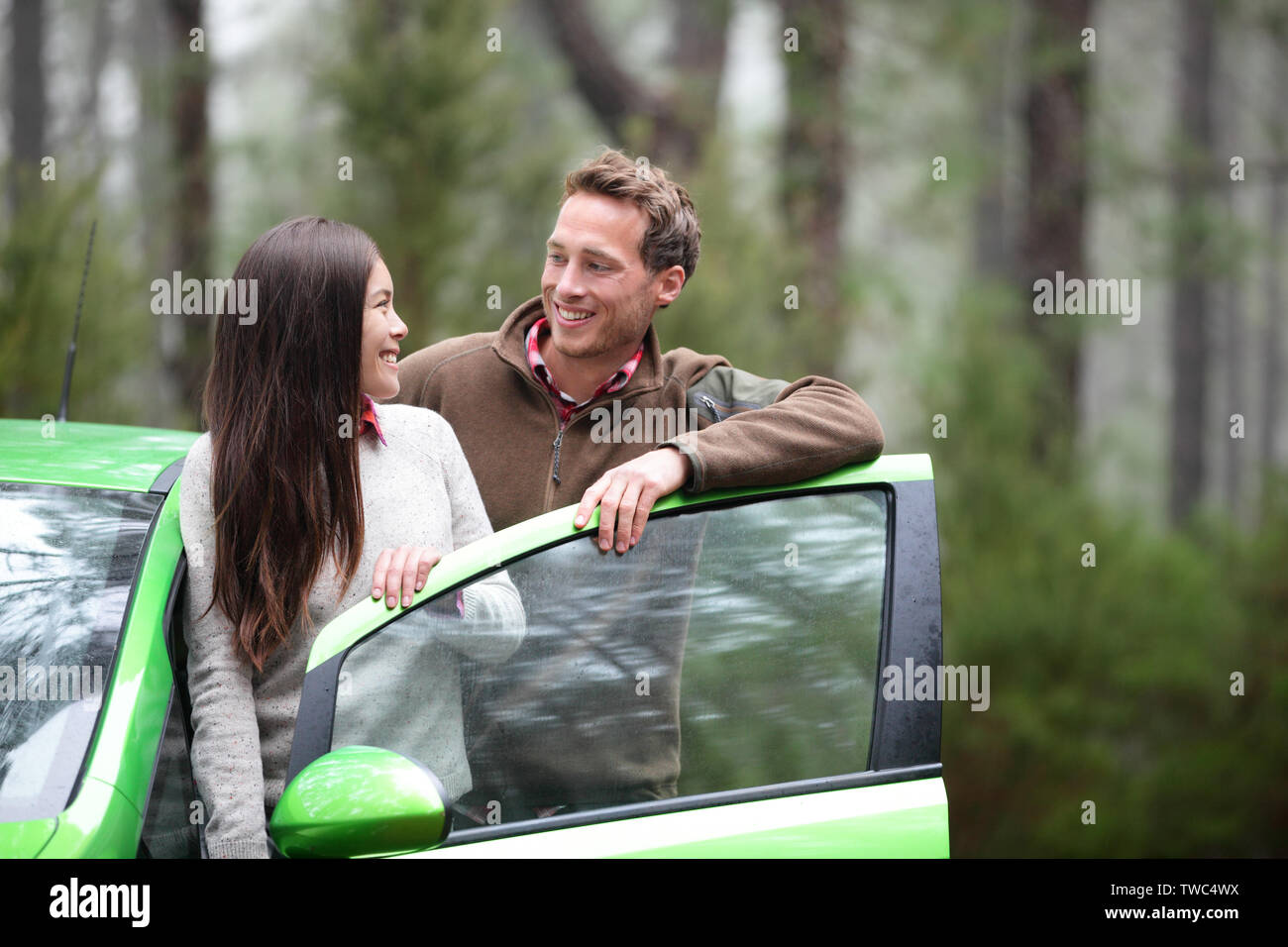 Car people - happy couple driving on road trip in green new rental car in beautiful nature forest. Romantic multiracial couple on the road on vacation. Asian woman, Caucasian man. Stock Photo